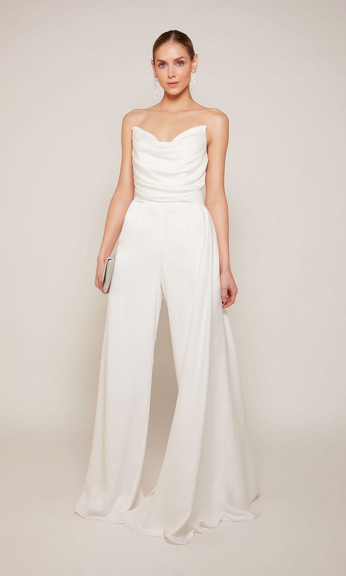 A woman poses against a neutral backdrop wearing the Long Formal Jumpsuit: Alyce 70050 in Diamond White, featuring wide legs and holding a small silver clutch.